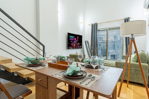 Bright living room with smart TV, green sofa and floor‑to‑ceiling windows overlooking street.