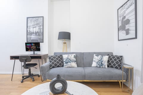 Living area with a plush gray futon accented with pillows, complemented by chic artwork and a modern floor lamp. A dedicated desk and chair provide a perfect spot for remote work or study, making this space both comfortable and functional for guests