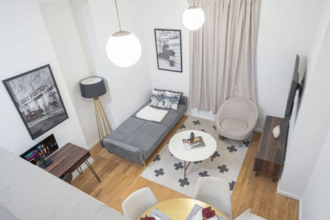 Bright loft view of the living area showcasing a versatile sofa bed, cozy armchair, and modern décor. The open layout flows seamlessly into the dining space below, highlighting wood floors, chic lighting, and tasteful artwork