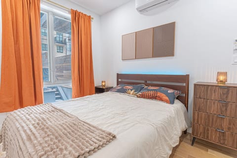 Cozy bedroom with a wooden bed, blue LED headboard light, and floral pillows. Features include bright orange curtains, a matching dresser, and three wall panels on white walls.