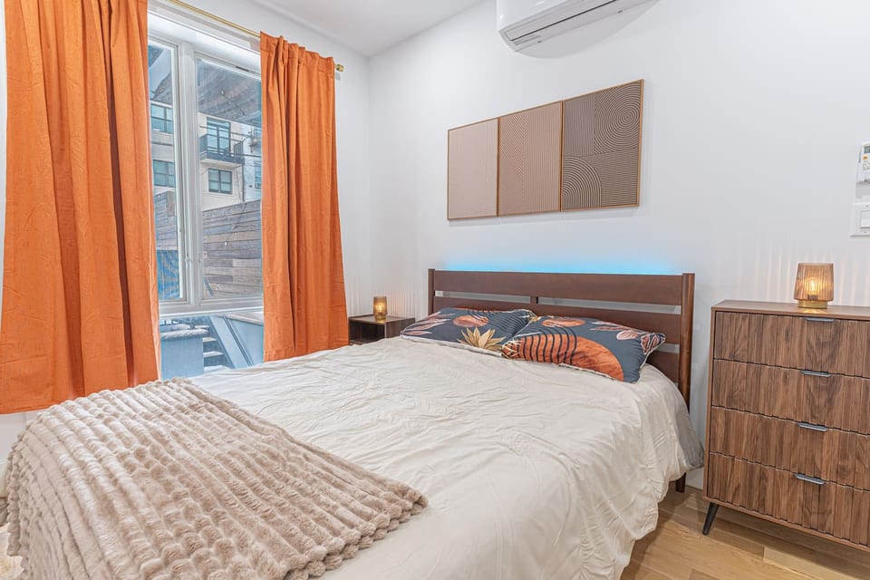 Cozy bedroom with a wooden bed, blue LED headboard light, and floral pillows. Features include bright orange curtains, a matching dresser, and three wall panels on white walls.