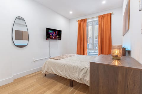 Bedroom with orange curtains, a wall-mounted TV, and an oval mirror. A wooden dresser with a glowing lamp sits in the table next to the bed.