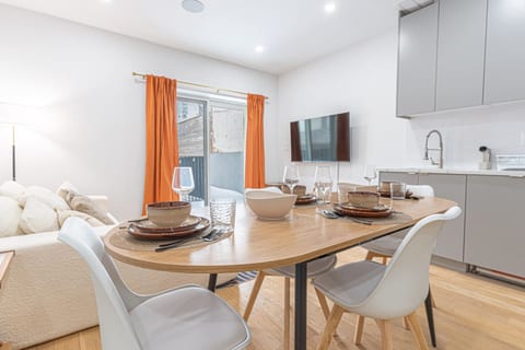 A set wooden dining table with utensils, white chairs in an open-concept living area. In the background, there are orange curtains, a TV, a white sofa, and kitchen cabinets.
