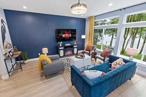 Bright waterfront living room with large windows, cozy fireplace & TV