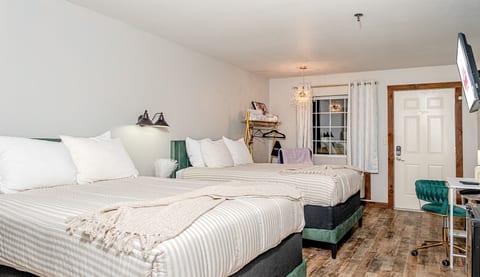 Clean double queen room with comfortable beds, bright natural light, workspace, in-room storage, and convenient access to the entrance.