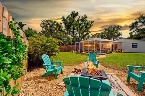 Golden-hour fire pit at Sun Oasis — teal Adirondack chairs, real wood flames, lush tropical palms, and a screened pool glowing in the background. The perfect Florida evening.