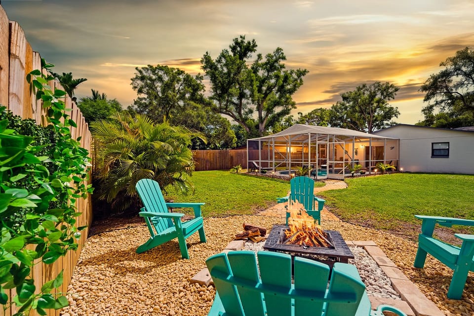 Golden-hour fire pit at Sun Oasis — teal Adirondack chairs, real wood flames, lush tropical palms, and a screened pool glowing in the background. The perfect Florida evening.