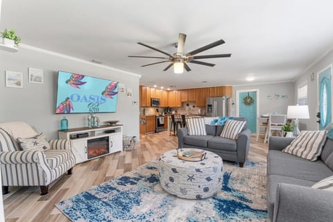 Open-concept living room at Sun Oasis with a large smart TV, fireplace credenza, coastal turtle artwork, beachy blue-and-white decor, and views straight through to the fully equipped kitchen.