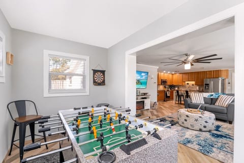 Game room open to the main living area at Sun Oasis — foosball table in the foreground, dartboard on the wall, and a seamless view through to the coastal living room and kitchen beyond.