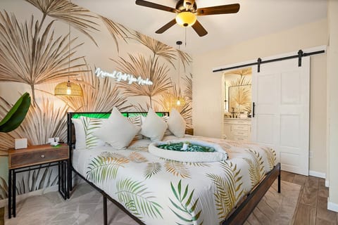 Primary king suite — palm-leaf mural, neon accent, woven pendant lights, and barn door entry to private spa ensuite. St. Petersburg, FL vacation rental.