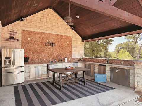Covered outdoor kitchen featuring full stainless steel refrigerator, expansive granite counters, full sink & bar on the side of the kitchen for open-air dining.