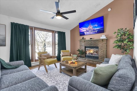 Spacious lakeview living room featuring two oversized couches, extra seating, a large TV, cozy fireplace, and ceiling fan—perfect for relaxing together after a day of fun.
