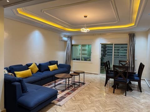 Spacious living room featuring a stunning golden tray ceiling with mood lighting, designer pendant lamp, plush blue corner sofa with yellow cushions, and direct sliding door access to the private terrace.