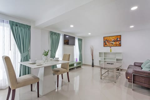 Bright dining area with a white table set for four, next to large windows with green curtains. Opens into the living space with a comfortable sofa and coffee table, making it ideal for meals and relaxation.