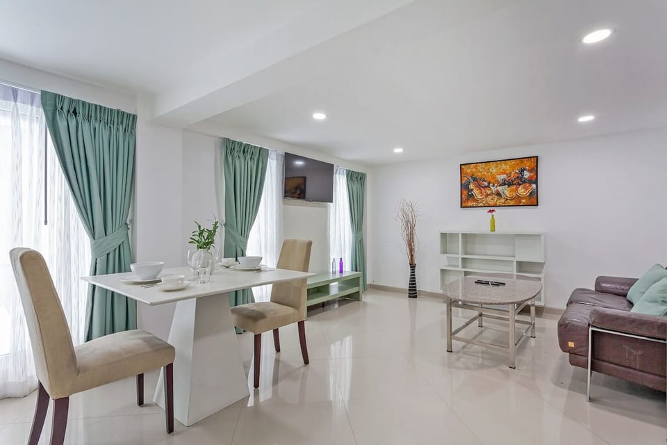 Bright dining area with a white table set for four, next to large windows with green curtains. Opens into the living space with a comfortable sofa and coffee table, making it ideal for meals and relaxation.