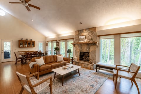 Bright and inviting living room with a cozy stone fireplace, vaulted ceilings, warm wood floors, comfortable seating, and beautiful forest views through expansive windows.