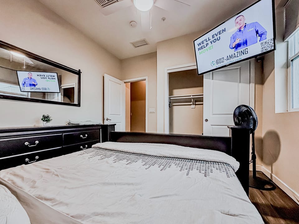 Bright bedroom with wall-mounted TV, dresser, open closet & ceiling fan.