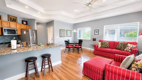 Bright open kitchen and living area with breakfast bar seating, wood-look flooring, cozy red seating, and plenty of natural light.