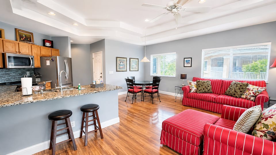 Bright open kitchen and living area with breakfast bar seating, wood-look flooring, cozy red seating, and plenty of natural light.