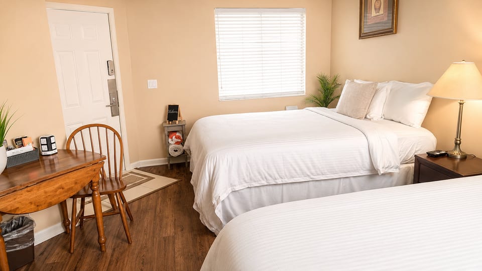 Spacious bedroom featuring two comfortable beds, small dining/work table, wood-look flooring, and plenty of natural light.