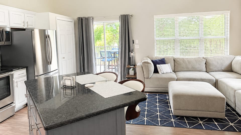 Updated open-concept kitchen and living area with stainless steel appliances, comfortable seating, and natural light from the balcony doors.