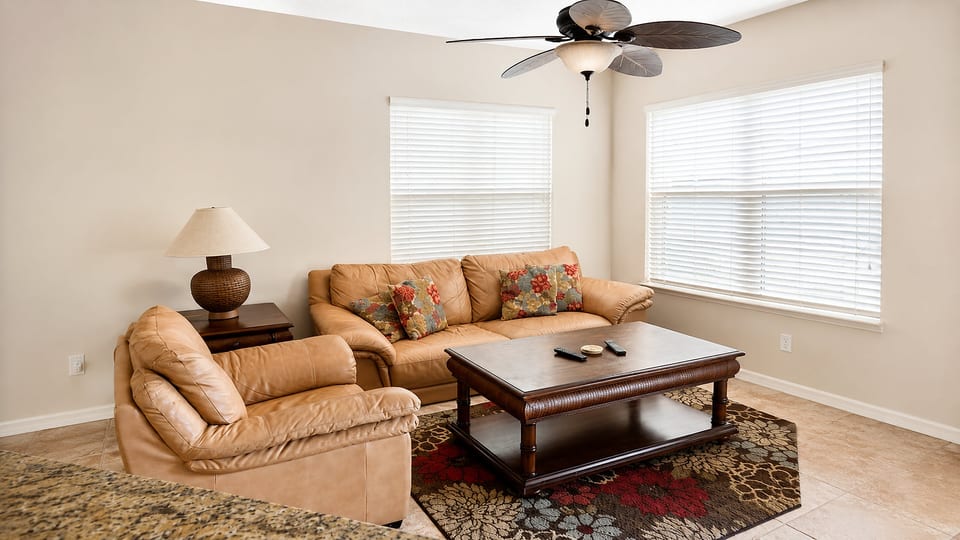 Comfortable living area with cozy sofa seating, ceiling fan, large windows, and a bright open feel.