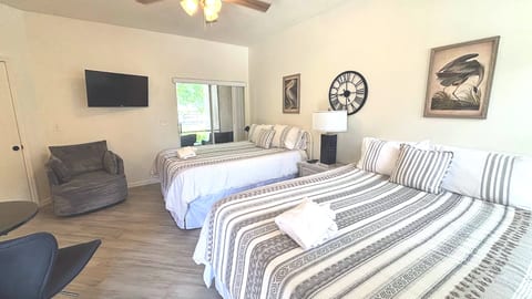 Bright bedroom featuring two beds, wall-mounted TV, cozy seating, and wood-look flooring.