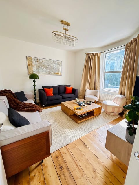 Light-filled living room featuring a 55" TV, plush seating, and a striking gold grid-mirror wall art piece. Complete with a statement chandelier, bay window, and rich wooden floors-ideal for relaxing or group movie nights.