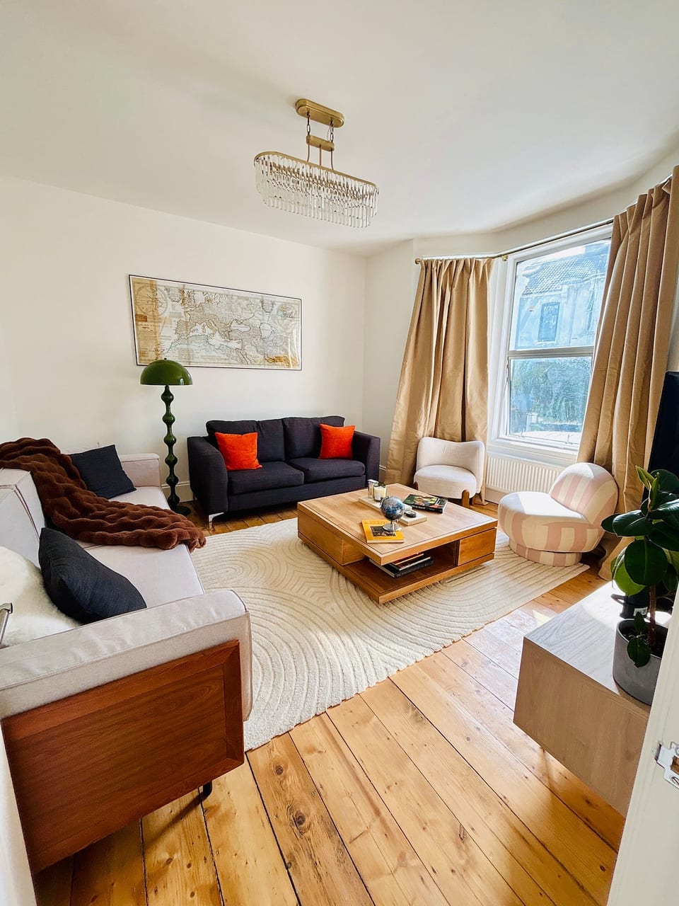Light-filled living room featuring a 55" TV, plush seating, and a striking gold grid-mirror wall art piece. Complete with a statement chandelier, bay window, and rich wooden floors-ideal for relaxing or group movie nights.