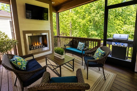 Screened outdoor patio surrounded by trees, featuring comfortable lounge seating, an outdoor fireplace, TV and gas grill—perfect for relaxing, entertaining or enjoying a quiet evening at the lake.