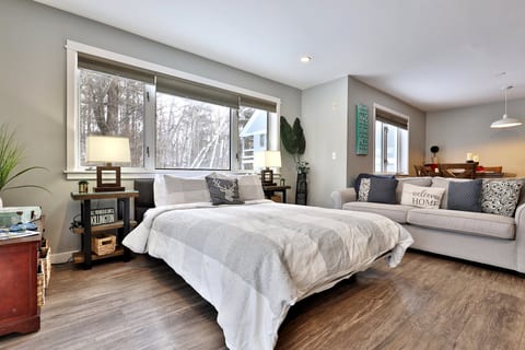 The queen bed sits between wide windows with the sectional sofa and dining area nearby.