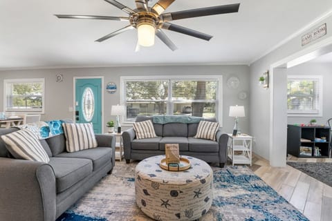 Full living room — two plush sofas, striped armchair, coastal round ottoman, oversized Oasis on 40th turtle 70-inch TV art, guest book on the coffee table, and the game room archway beyond.