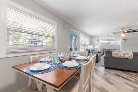 Coastal dining room at Sun Oasis — a farmhouse-style table set for six with blue place settings and teal glassware, teal front door visible beyond, flowing into the living room.