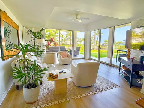 Bright living room with ecru seating, tropical plants, large sliding glass doors, and views of lanai, landscaped grounds, ocean views. Open-concept space, comfortable seating, TV, abundant natural light, and easy access to outdoor living areas.