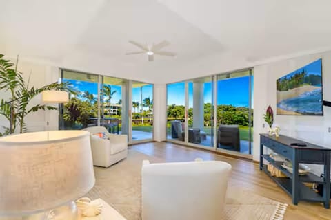 Bright living room with ecru seating, tropical plants, large sliding glass doors, and views of lanai, landscaped grounds, ocean views. Open-concept space, comfortable seating, TV, abundant natural light, and easy access to outdoor living areas.