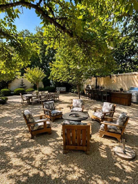 Inviting outdoor gathering space shaded by mature trees, featuring comfortable seating around a fire pit, string lights, and an outdoor kitchen with a grill and bar seating—perfect for relaxing and entertaining.