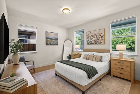 A bright and inviting queen bedroom with stylish furnishings, warm wood tones, and abundant natural light. A Smart TV and comfortable seating create a relaxing space to unwind.