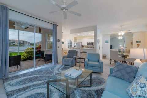 A bright, modern living room with comfortable seating, a plush area rug, and a glass coffee table. Large glass doors open to a screened lanai, creating a seamless indoor-outdoor feel.