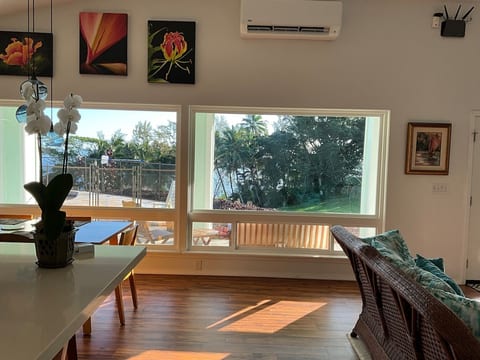 View towards large dining room windows with pool, palm trees and ocean in the background. To the right is the wicker couch. On the left is the edge of the kitchen island. And on the walls is the AC unit and various pieces of local art.
