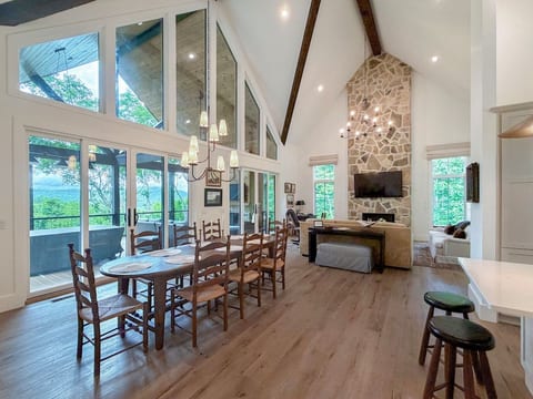 Welcoming dining area designed for gathering, overlooking the spacious great room with soaring chalet ceilings, a cozy fireplace, oversized windows, and beautiful mountain views beyond the glass.