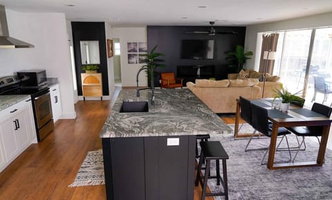 Bright, modern living space featuring a granite kitchen island, stainless appliances, dining seating, and a comfortable living area with TV for relaxing after the pool.