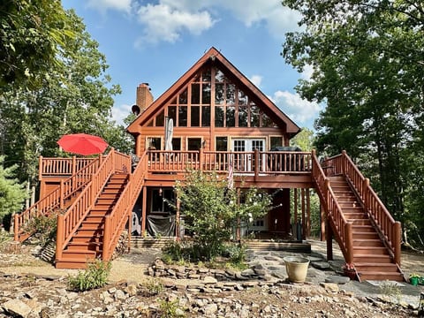Wooden deck with log railing all around including a side deck with sun beds and a hammock for two people. Outdoor dining set and lounge beds with two patio umbrellas and a professional gas grill.