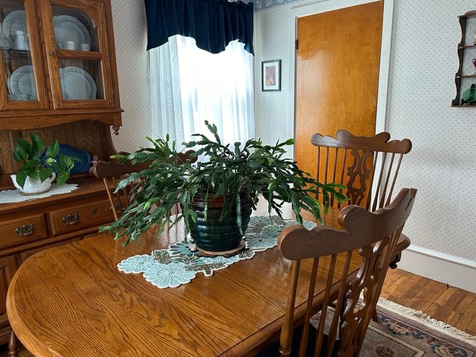 Enjoy a cozy dining room with a classic wooden table and chairs, perfect for meals and gatherings. The space is brightened by natural light and accented with houseplants and vintage decor, offering a warm, inviting atmosphere.