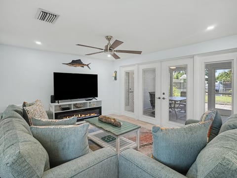 Relax in the bright, coastal-inspired living room featuring a spacious sectional, large-screen TV, electric fireplace, and direct access to the screened lanai.