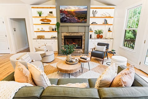 The main living area features vaulted ceilings, exposed wood beams, a 65" Smart TV above the stone fireplace, and built-in shelving. Plenty of seating for the whole group.