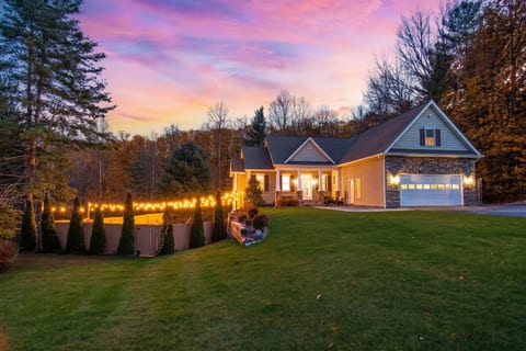 The front of Haven in the Hills at sunset. Modern farmhouse style on a quiet, wooded lot with string lights lining the pool fence and a covered front porch.