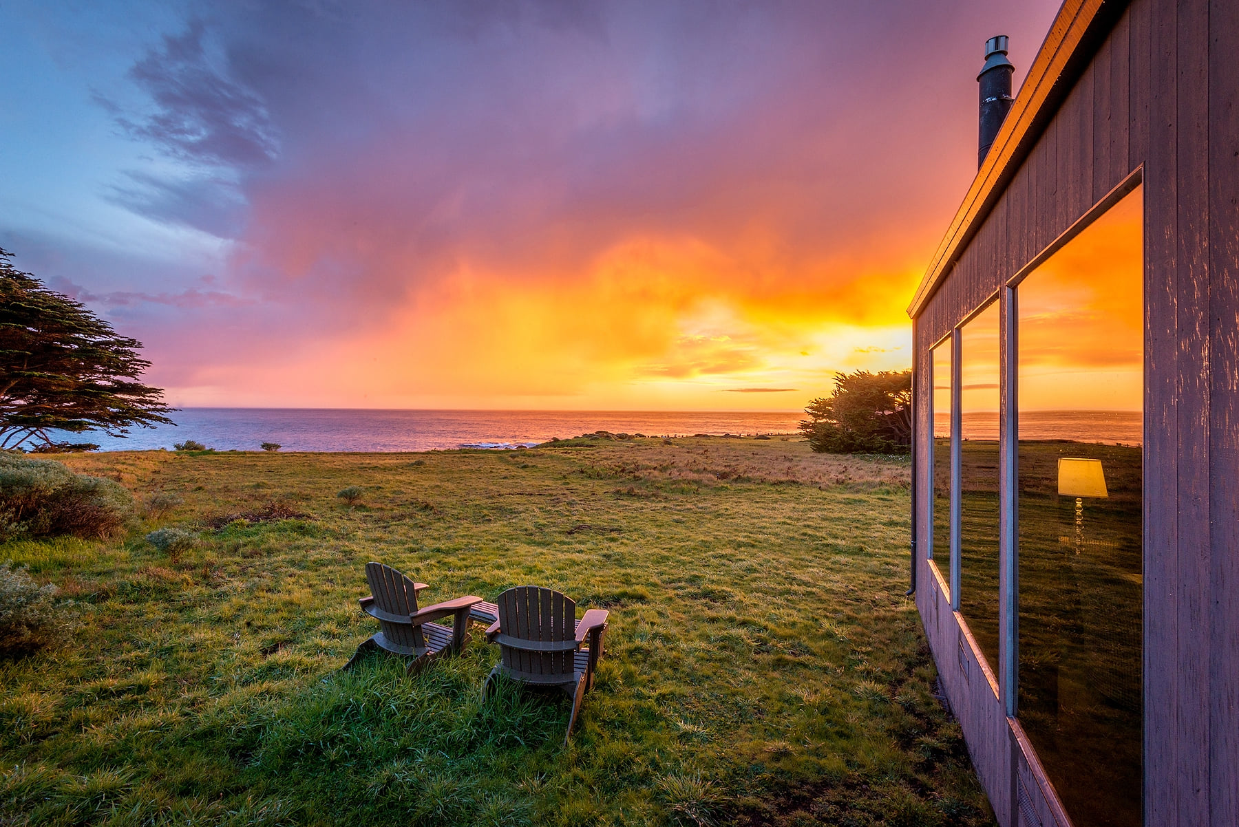 Luxury oceanfront escape: Two Adirondack chairs overlook a glowing orange sherbet colored sunset on the Pacific, reflected in the sleek windows of a modern coastal retreat—pure serenity, steps