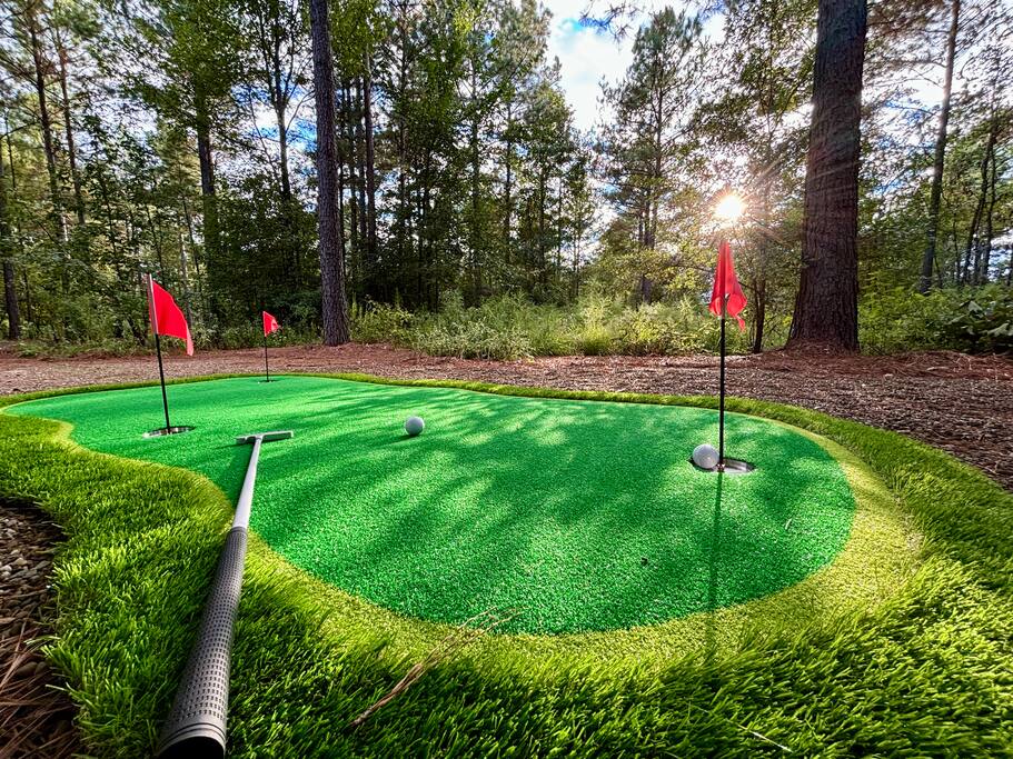 Serene outdoor putting green with red flags and golf gear under a sunlit canopy of trees.