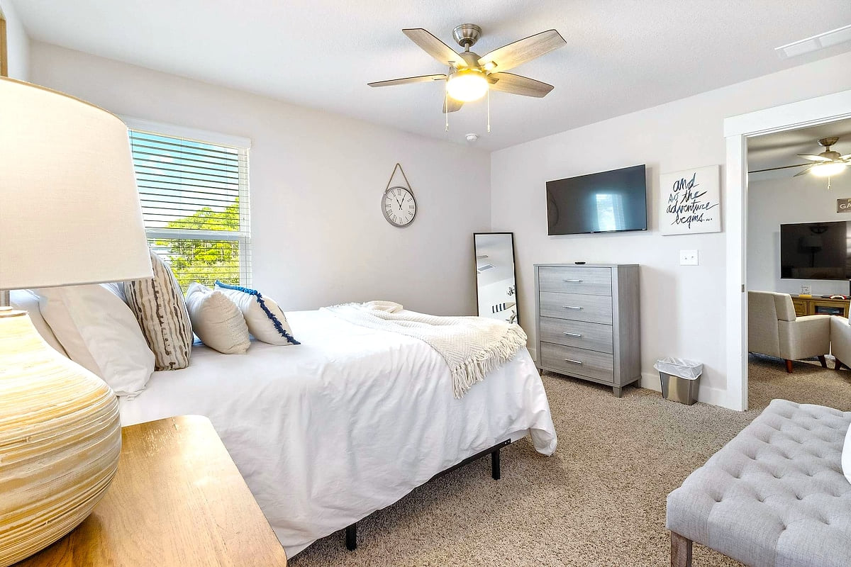 Sunny bedroom with queen bed, smart TV, fan, and full-length mirror.