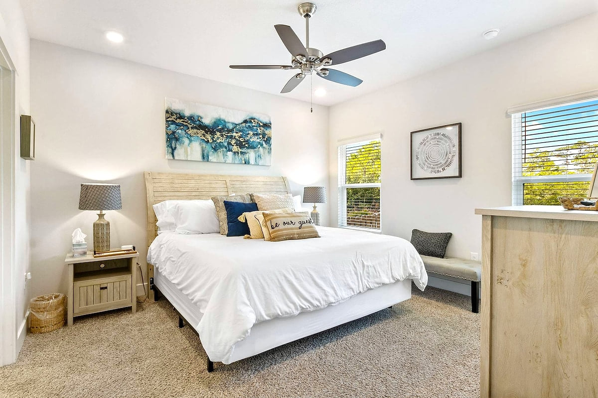Bright king suite with coastal accents, plush carpet, fan, and sunlit windows.